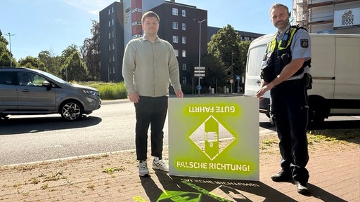 Zwei Männer halten eine grünbesprühte Schablone. Darauf steht "Falsche Richtung!" und spiegelverkehrt "Gute Fahrt!"