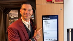 Der Zugchef steht imn Zug und zeigt lächelnd auf eine Anzeige mit Zielbahnhof Ostende