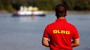 Ein Mitarbeiter der DLRG steht am Rheinufer, im Hintergrund ein Boot der Wasserschutzpolizei