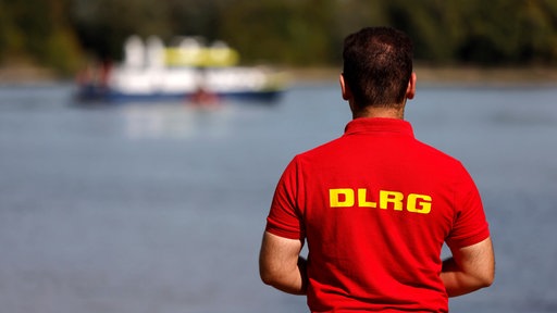 Ein Mitarbeiter der DLRG steht am Rheinufer, im Hintergrund ein Boot der Wasserschutzpolizei