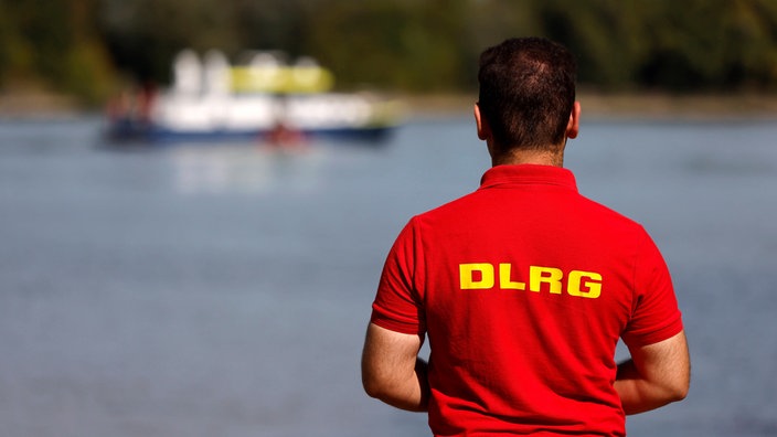 Ein Mitarbeiter der DLRG steht am Rheinufer, im Hintergrund ein Boot der Wasserschutzpolizei