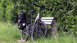Ein schwarzes Fahrrad mit einer Fahrradtasche steht an einer Bank angelehnt.