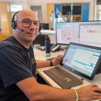 Ein Mann sitzt in einem Büroraum vor Computerbildschirmen und hat ein Headset auf.