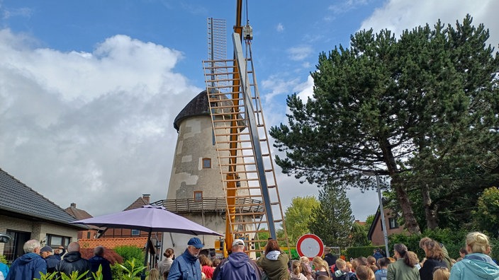 Ein Kran setzt neue Flügel an eine Mühle, Zuschauer stehen davor