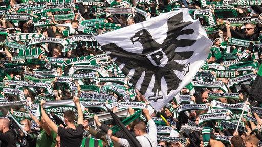 Fans von Preußen Münster halten Schals und Flaggen des Vereins in die Luft. 