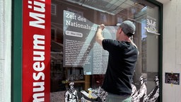 Mann klebt großen Aufkleber and Fensterscheibe mit einem Infotext "Zeit des Nationalsozialismus" 