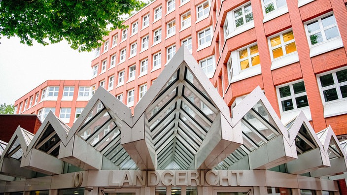 Ein gezacktes Vordach aus Metall und Glas, darunter der Schriftzug "Landgericht" über einer Schiebetür. Im Hintergrund ragt ein mehrstöckiges Bürohaus aus rotem Backstein hervor.