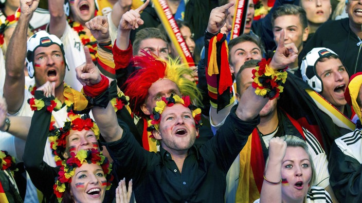 Ausschnitt von einer jubelnden Menge an Menschen beim Public Viewing. Alle tragen deutsche Fußball-Fanartikel wie Fußballtrikots oder Blumenketten in schwarz, rot, gelb.