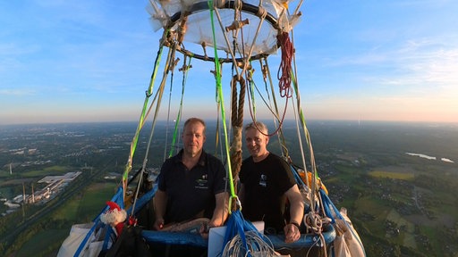 Ballonfahrer Andreas Zumrode und Axel Hunnekuhl stehen in dem Korb eines Gasballons, der in der Luft fliegt. Im Hintergrund ist eine große grüne Landschaft und einzelne Häuser von oben zu erkennen.