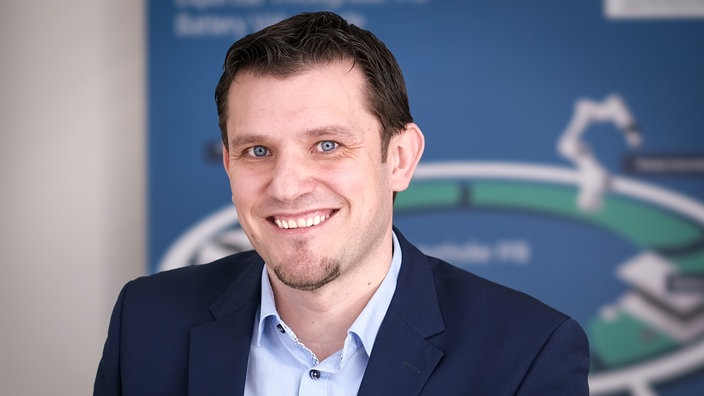 Über Batteriespeicher in NRW: Ein Mann mit kurzen Haaren und einem Anzug vor einem blau-weißen Hintergrund