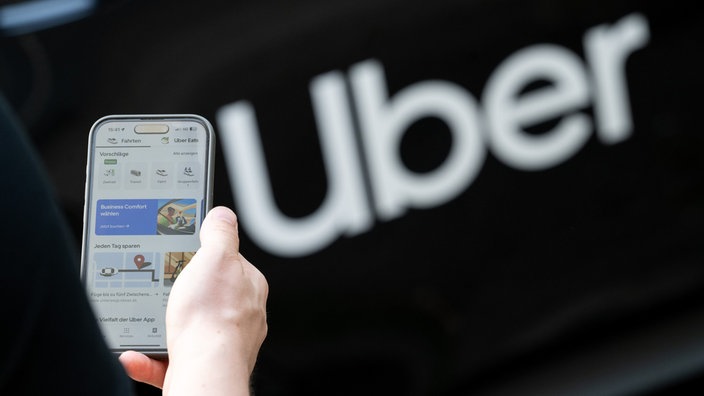 Jemand hält ein Smartphone mit der Uber-App in der Hand, im Hintergrund der Schriftzug des Unternehmens.
