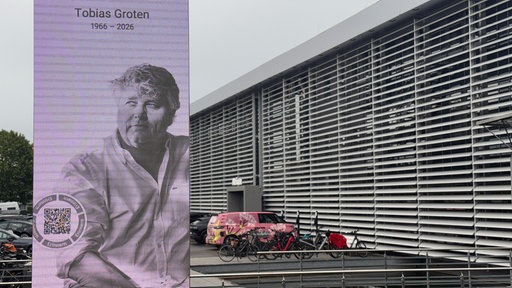Bildschirm mit Bild von Tobias Groten mit Lebensdaten vor dem Tobit Gebäude.