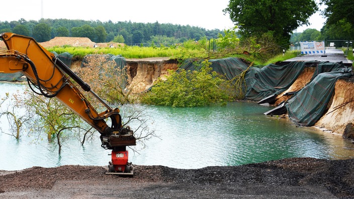 Abrisskante an einem See nach einem Erdrutsch, im Vordergrund der Arm einer Baumaschine.