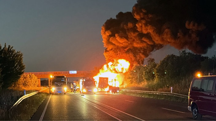Ein brennedes Fahrzeug mit großer Rauchwolke steht auf dem Seitenstreifen. Daneben steht die Feuerwehr.