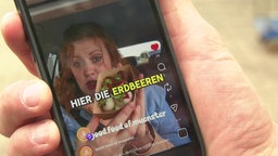 Handy in Hand, darauf ein Video, in dem eine Frau Erdbeeren in die Kamera hält