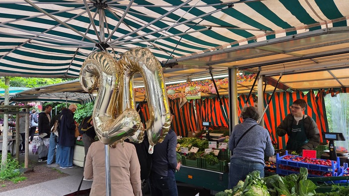 Stand auf einem Bauernmarkt. Luftballons in Form einer 20. Menschen kaufen auf dem Markt ein.