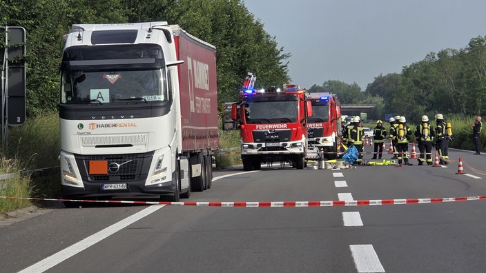 Links ist ein LKW, dahinter zwei Feuerwehrfahrzeuge. Rechts stehen einige Feuerwehrmänner in Montur.