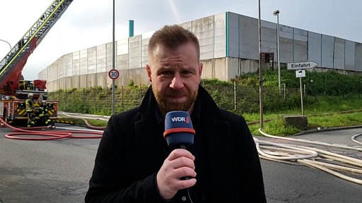 WDR-Reporter Johannes Hoppe steht mit einem Mikrofon in der Hand vor einer großen Halle, im Hintergrund arbeitet die Feuerwehr