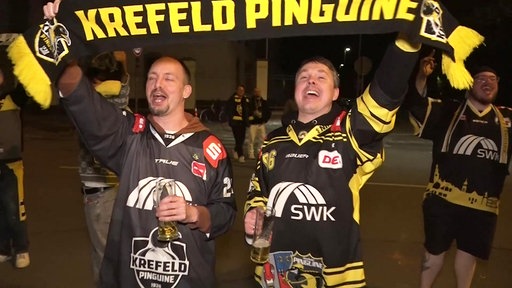 Zwei Fans des Eishockey-Clubs Krefeld Pinguine in Mannschaftstrikots jubeln ausgelassen und halten einen schwarz-gelben Fanschal mit dem Vereinsnamen hoch.