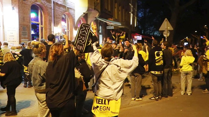 Fünfzig Fans des Eishockey-Clubs Krefeld Innenstadt tragen Kleidung in den Vereinsfarben schwarz und gelb und feiern ausgelassen vor einer Gaststätte.