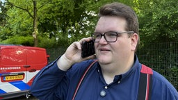 Ein Mann mit Brille hält sich sein Handy ans Ohr. Im Hintergrund ein Fahrzeug der Brandweer.