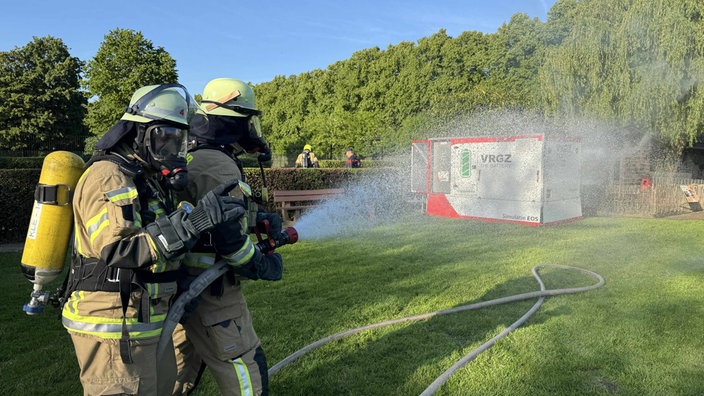 Zwei Feuerwehrleute in Schutzkleidung richten einen Löschschlauch auf einen brennenden Generator 