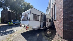 Ein weißer Wohnwagen steht schräg vor einer Hauswand. Die vordere Kante ist zerbeult. Auf der anderen Straßenseite steht ein Polizeiauto.