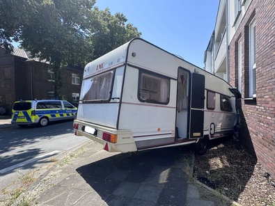 Ein weißer Wohnwagen steht schräg vor einer Hauswand. Die vordere Kante ist zerbeult. Auf der anderen Straßenseite steht ein Polizeiauto.