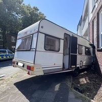 Ein weißer Wohnwagen steht schräg vor einer Hauswand. Die vordere Kante ist zerbeult. Auf der anderen Straßenseite steht ein Polizeiauto.