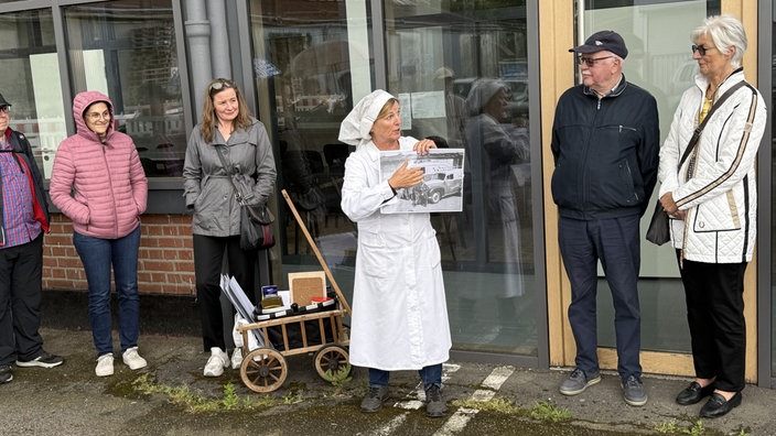 Eine Dame in einer Bäckeruniform hält ein Bild hoch und zeigt darauf, während sie weitere Personen anschauen.