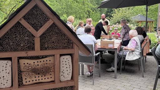 Im Vordergrund ist ein großes Insektenhotel zusehen, dahinter arbeiten Menschen am Tisch an einem weiteren Insektenhotel