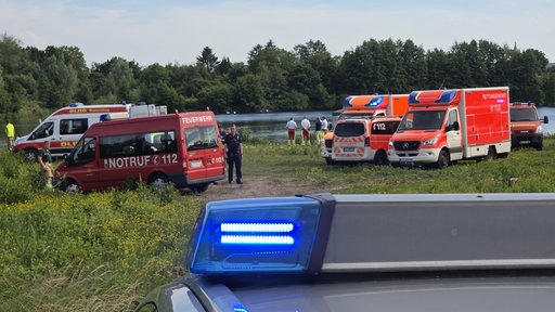 Im Hintergrund ist ein See, davor stehen einige Menschen in Rettungs-Uniformen. Rechts daneben stehen mehrere Krankenwagen und ein Notarztwagen. Links im Bild sind noch mehr Rettungsfahrzeuge. Im Vordergrund sieht man das Dach eines Polizeiautos.
