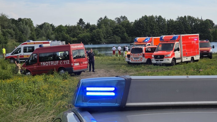 Im Hintergrund ist ein See, davor stehen einige Menschen in Rettungs-Uniformen. Rechts daneben stehen mehrere Krankenwagen und ein Notarztwagen. Links im Bild sind noch mehr Rettungsfahrzeuge. Im Vordergrund sieht man das Dach eines Polizeiautos.