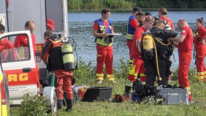 Taucher und Rettungskräfte der DLRG stehen auf einer Wiese. Im Hintergrund ein See.