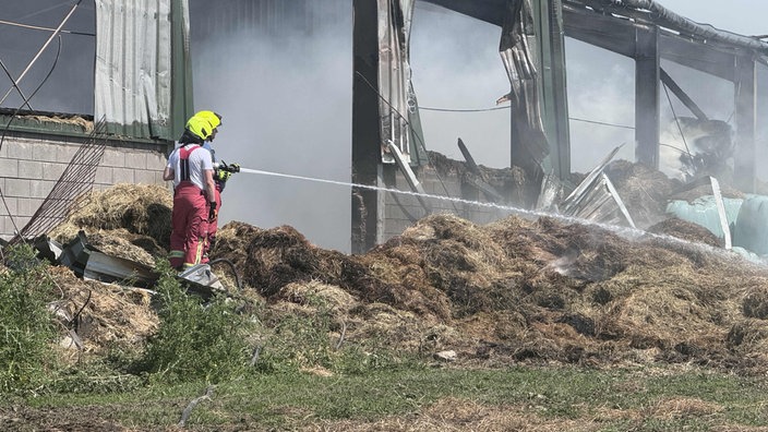Zwei Feuerwehrleute in Einsatzkleidung und mit gelben Helmen spritzen Waser auf Stroh. Dahinter ist eine Scheune mit verkohltem Blech.