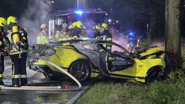 Ein gelber Mustang, der gegen einen Baum gekracht ist. Der Mustang ist ausgebrannt. Dahinter stehen Feuerwehrmänner und ein Feuerwehrauto.