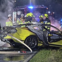 Ein gelber Mustang, der gegen einen Baum gekracht ist. Der Mustang ist ausgebrannt. Dahinter stehen Feuerwehrmänner und ein Feuerwehrauto.