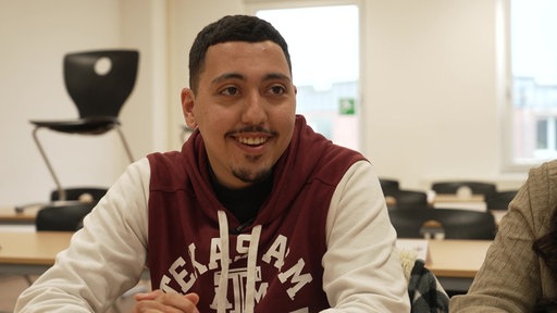 Youssef lächelt in die Kamera. Er trägt einen rot-weißen Pullover und hat die Hände verschränkt.