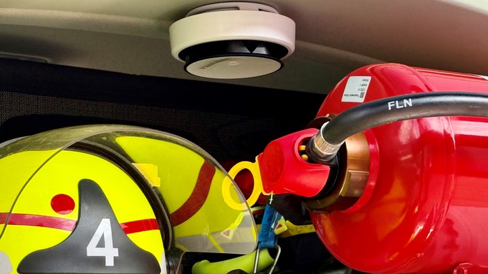 An der Decke eines Feuerwehrautos häbft ein weißer, runder Feuermelder. davor ein roter Feuerlöscher und ein Helm.