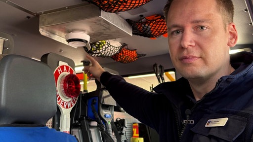 Der freiwillige Feuerwehrmann Marc Vinschen zeigt auf einen Rauchmelder, der innen an der Decke eines Einsatzfahrzeuges hängt.