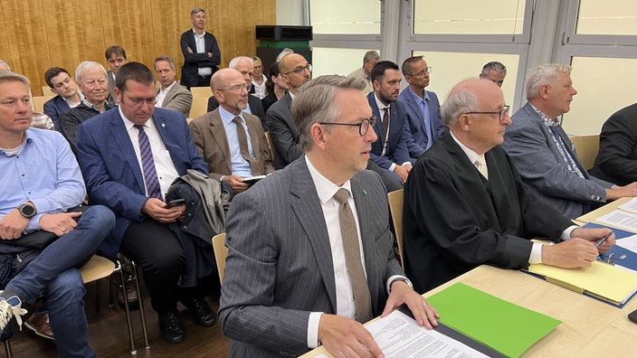 Landrat Ingo Brohl im Gerichtssaal. Er trägt einen grauen Anzug und eine braune Krawatte. Hinter ihm sitzen noch weitere Männer in Anzügen.