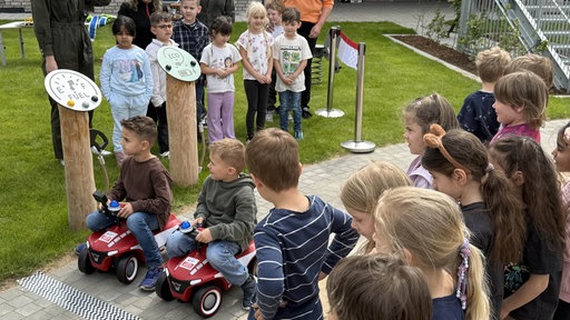 Zwei Jungen sitzen auf Bobbycars und weitere Kinder beobachten die beiden.