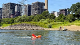 Person hält sich im Rhein an einem orangefarbenen Wasserrettungsroboter fest. Im Hintergrund sind das Rheinufer, ein Rettungsschwimmer auf einem Steg sowie Industrieanlagen und Hochspannungsleitungen zu sehen.