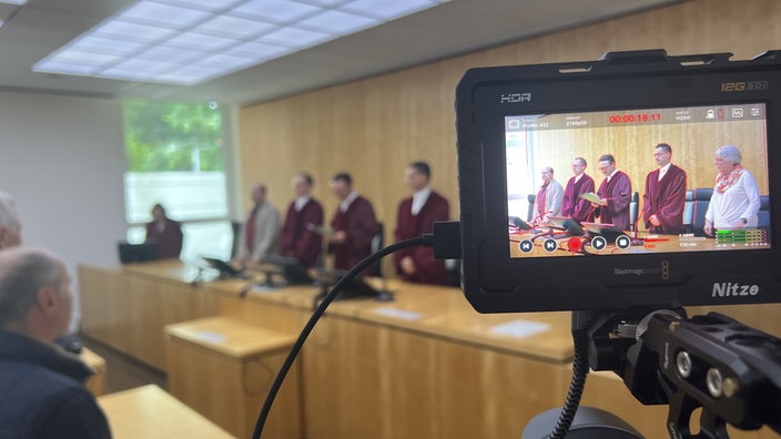 Im Hintergrund stehen die Richter vor Tischen. Der Bildausschnitt ist unscharf. Vorne rechts sieht man das Bild durch einen Kamerabildschirm in scharf.