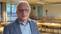 Ein älterer Mann im Anzug mit weißen Haaren und Brille schaut in die Kamera. Er steht in einem Kirchenraum mit mehreren Tischen und Stühlen, an der Wand hängt ein Kreuz.