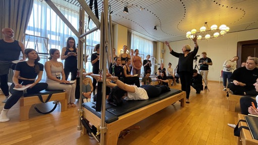 Eine Trainerin erklärt eine Übung an einem Pilates Gerät, während TeilnehmerInnen drumherum stehen und zuschauen