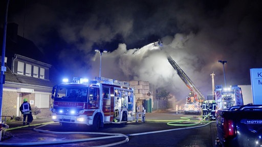 Einsatzfahrzeuge der Feuerwehr, inklusive einer Drehleiter, stehen am Gelände einen Müllentsorgers. Im Hintergrund befindet sich eine große Rauchwolke.