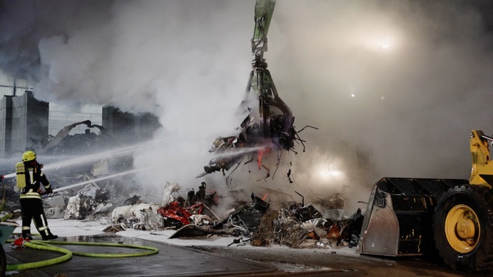 Einsatzkräfte der Feuerwehr zielen mit Wasserstrahlen auf einen glühenden Schrotthaufen, der von einem Greifarm gehalten wird. Rundherum befindet sich dichter Rauch.