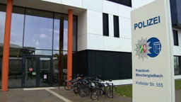 Der Eingang zum Polizeipräsidium Mönchengladbach: ein hohes Glasfoyer mit zwei orangen Säulen in einem mit schwarzen und weißen Platten verkleideten Flachbau, davor eine Stele mit dem Logo der Polizei.