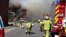 Einsatzwagen der Feuerwehr und Einsatzkräfte stehen vor einer großen Rauchwolke. 